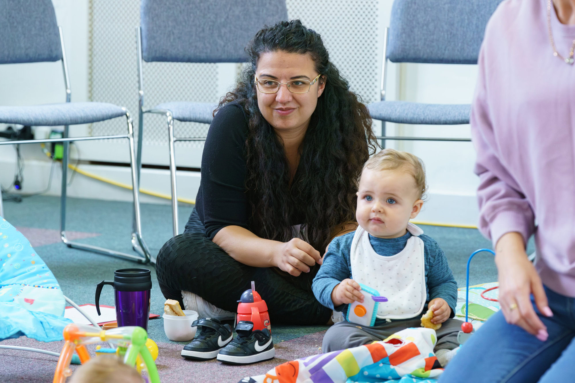 Woman sitting on the floor behind a baby wearing a bib and playing with toys